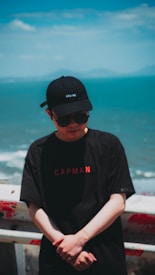 A person wearing a black cap and sunglasses stands in front of a blue ocean with a partly cloudy sky. They are wearing a black t-shirt with red lettering and are standing against a railing that has red marks.