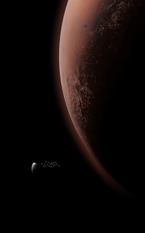 A reddish-brown planet dominates the right side of the image, showing detailed surface textures and subtle color variations. A small, grayish celestial body, possibly a moon, is visible to the left against a black space backdrop.