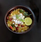 A bowl of poha garnished with grated coconut, peanuts, curry leaves, a lemon wedge, and cilantro is placed against a dark background.