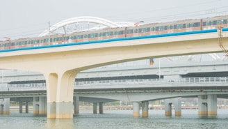 A train travels along an elevated railway bridge above water, with multiple levels of infrastructure visible. The bridge is supported by large concrete pillars, and the train's colorful exterior contrasts with the muted tones of the surrounding structures.