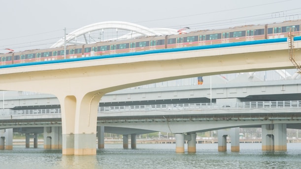 A train travels along an elevated railway bridge above water, with multiple levels of infrastructure visible. The bridge is supported by large concrete pillars, and the train's colorful exterior contrasts with the muted tones of the surrounding structures.