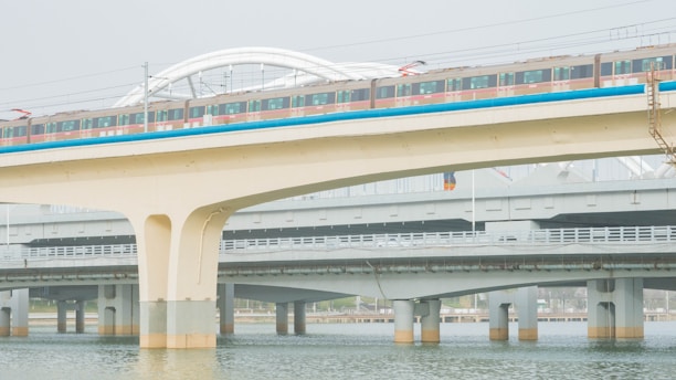 A train travels along an elevated railway bridge above water, with multiple levels of infrastructure visible. The bridge is supported by large concrete pillars, and the train's colorful exterior contrasts with the muted tones of the surrounding structures.