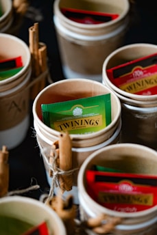 A collection of small cups hold assorted tea bags, including a prominent green and yellow Twinings packet. Some cups are tied with twine and sticks of cinnamon are placed alongside the packets, enhancing the rustic presentation.