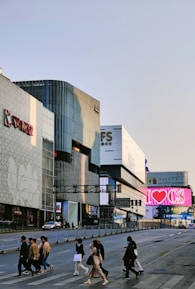 A bustling urban street scene with several people crossing at a crosswalk. Tall modern buildings with large advertisements and shop signs line the street, including a prominent 'I ♥ CS' billboard. The environment appears busy with both pedestrian and vehicle traffic.