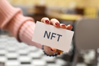 A hand with red nail polish holds a white card with the text 'NFT' written on it. The background is blurred, featuring black and white checkered flooring and beige-colored furniture.
