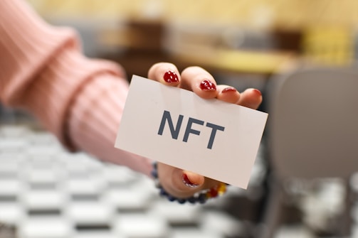 A hand with red nail polish holds a white card with the text 'NFT' written on it. The background is blurred, featuring black and white checkered flooring and beige-colored furniture.