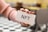 A hand with red nail polish holds a white card with the text 'NFT' written on it. The background is blurred, featuring black and white checkered flooring and beige-colored furniture.