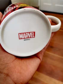A close-up view of the underside of a white ceramic mug being held by a person. The bottom of the mug has a red and white Marvel logo with a small copyright symbol beneath it. The background has a wood-patterned floor and part of a white door.