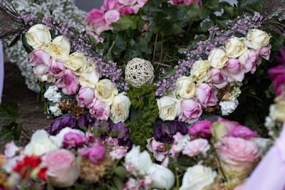 A detailed arrangement of flowers featuring white and pink roses, baby's breath, and green foliage. A central spherical object made of woven material is surrounded by the blooms, creating an intricate floral design. The composition includes varying shades of purple along with textured greenery.