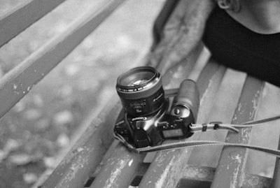 A black and white photo captures a digital camera resting on a worn wooden park bench. A person's arm is partially visible, suggesting casual interaction or rest. The camera is equipped with a lens and is tethered with a strap.