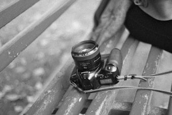 A black and white photo captures a digital camera resting on a worn wooden park bench. A person's arm is partially visible, suggesting casual interaction or rest. The camera is equipped with a lens and is tethered with a strap.