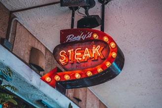 A retro-style illuminated sign with neon lettering that reads 'Steak Here!' and is adorned with a border of light bulbs. The sign is predominantly red with a directional arrow shape pointing downward. It hangs against a textured wall, with some greenery visible around the edges.