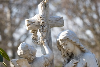 A stone sculpture depicting an angel embracing a cross adorned with flowers, alongside a woman with a serene expression. The scene conveys a sense of peace amidst a backdrop of blurred trees.