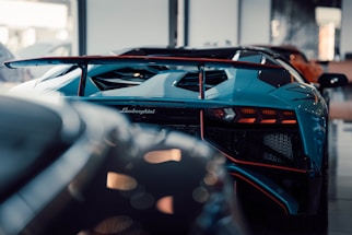 A close-up view of the rear of a sleek, blue Lamborghini sports car with an aerodynamic design. The focus is on the car's intricate rear details, including the spoiler, tail lights, and the Lamborghini logo. The setting appears to be indoors, possibly a showroom, with other cars faintly visible in the background.