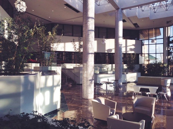 A spacious, modern lobby area featuring large windows allowing ample natural light to flood the space. The lobby is decorated with multiple indoor plants, round tables surrounded by chairs, and contemporary armchairs. Tall columns contribute to the openness, with glossy floors reflecting the ambient light.