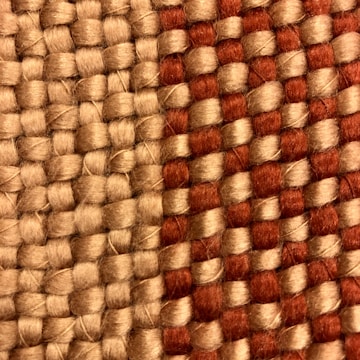 A close-up view of a woven fabric featuring interlocking strands of beige and reddish-brown threads. The texture of the fabric is coarse, highlighting the thick yarns used in the weaving process.