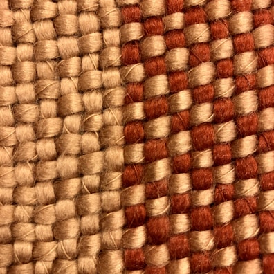 A close-up view of a woven fabric featuring interlocking strands of beige and reddish-brown threads. The texture of the fabric is coarse, highlighting the thick yarns used in the weaving process.