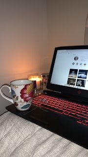A cozy setup featuring a laptop with a glowing red keyboard, opened to a webpage. A floral-patterned mug sits to the left, and a lit candle adds a warm ambiance to the scene.