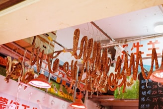 Various cured meat sausages hang from hooks under a ceiling adorned with bright red heating lamps. The background includes vibrant signage with colorful writing, and a blackboard on the side displays prices. The overall setting suggests a market or butcher shop, giving a sense of bustling commerce.