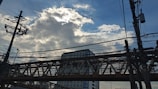 A network of electrical wires and utility poles is silhouetted against a partly cloudy sky. A tall building and an overpass are visible in the background, with the sun partially obscured by clouds, creating a striking interplay of light and shadow.