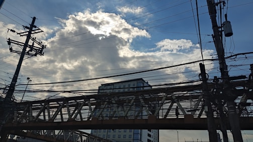 A network of electrical wires and utility poles is silhouetted against a partly cloudy sky. A tall building and an overpass are visible in the background, with the sun partially obscured by clouds, creating a striking interplay of light and shadow.