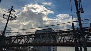 A network of electrical wires and utility poles is silhouetted against a partly cloudy sky. A tall building and an overpass are visible in the background, with the sun partially obscured by clouds, creating a striking interplay of light and shadow.