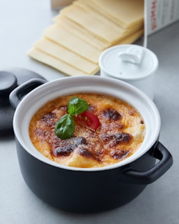 A ceramic pot filled with a baked dish topped with browned cheese, garnished with a basil leaf and a slice of tomato. In the background, there are pieces of lasagna sheets and a small white container.