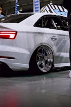 A sleek white car is showcased indoors, featuring large chrome wheels and a polished body. The vehicle is positioned on a concrete floor with reflections visible on its surface. In the background, banners and signage with a blue theme can be seen. People are partially visible, suggesting the setting is an automotive event or exhibition.