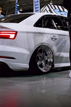 A sleek white car is showcased indoors, featuring large chrome wheels and a polished body. The vehicle is positioned on a concrete floor with reflections visible on its surface. In the background, banners and signage with a blue theme can be seen. People are partially visible, suggesting the setting is an automotive event or exhibition.