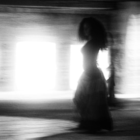 A monochrome, blurred image captures a person in motion, against a bright light source. The scene creates a mystical and enigmatic atmosphere with the figure's silhouette appearing somewhat abstract.