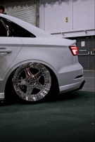 A white car with detailed shining chrome wheels is parked indoors, showcasing a sleek modern design. The car appears to be lowered, with minimal clearance between the fender and the tire. The background consists of a partially visible roller door and some industrial elements.
