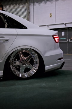 A white car with detailed shining chrome wheels is parked indoors, showcasing a sleek modern design. The car appears to be lowered, with minimal clearance between the fender and the tire. The background consists of a partially visible roller door and some industrial elements.