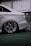 A white car with detailed shining chrome wheels is parked indoors, showcasing a sleek modern design. The car appears to be lowered, with minimal clearance between the fender and the tire. The background consists of a partially visible roller door and some industrial elements.