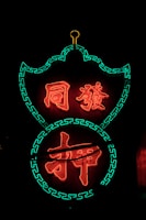 Bright neon sign with Chinese characters illuminated in red, outlined by green decorative patterns against a dark background.
