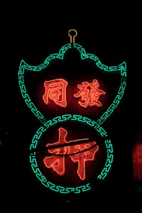 Bright neon sign with Chinese characters illuminated in red, outlined by green decorative patterns against a dark background.