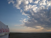 A wide open sky filled with scattered, fluffy clouds casting various shades of gray and white as the sun sets or rises, creating a serene atmosphere. A yurt with decorative patterns is partially visible on the left side, positioned on a grassy expanse leading to a calm body of water in the distance. Small figures of people can be seen near the water, enhancing the sense of scale and tranquility.
