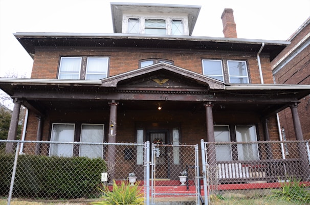 A large, two-story brown brick house with multiple windows, a central front door, and a covered front porch supported by columns. A chain-link fence surrounds the property, and there are a few plants and shrubs in the front yard. The house has a classic, somewhat aged appearance.