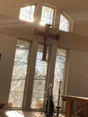 Sunlight filters through tall stained glass windows, casting rays of light across a serene church interior. A large wooden cross is mounted prominently on the wall, illuminated by the natural light. Elegant candlesticks and plants are arranged near the altar, contributing to the peaceful atmosphere.