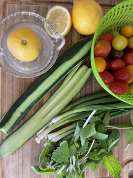 A variety of fresh vegetables and fruits are arranged on a wooden surface. There is a lemon in a glass juicer, a whole lemon, a sliced half lemon, several cucumbers, spring onions, mint leaves, and a green plastic basket containing assorted cherry tomatoes of different colors.