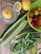 A variety of fresh vegetables and fruits are arranged on a wooden surface. There is a lemon in a glass juicer, a whole lemon, a sliced half lemon, several cucumbers, spring onions, mint leaves, and a green plastic basket containing assorted cherry tomatoes of different colors.