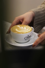 A person's hands, with neatly manicured nails, are holding a white cup of coffee with latte art on top. The cup sits on a white saucer and has 'Story Coffee' written on it.