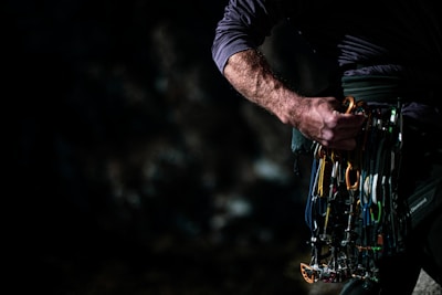 A person is holding a collection of climbing gear, specifically carabiners and other climbing equipment, against a dark background. The scene appears to be in low light, focusing on the hand and gear.