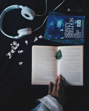 A pair of white headphones lies on a dark surface next to an open book. The book, surrounded by scattered flower petals, is being held open by a hand. Beside the book, there is another closed book titled 'Sidney Sheldon The Naked Face.' A single green leaf rests on the open pages.