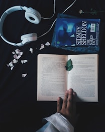A pair of white headphones lies on a dark surface next to an open book. The book, surrounded by scattered flower petals, is being held open by a hand. Beside the book, there is another closed book titled 'Sidney Sheldon The Naked Face.' A single green leaf rests on the open pages.