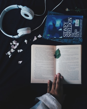 A pair of white headphones lies on a dark surface next to an open book. The book, surrounded by scattered flower petals, is being held open by a hand. Beside the book, there is another closed book titled 'Sidney Sheldon The Naked Face.' A single green leaf rests on the open pages.