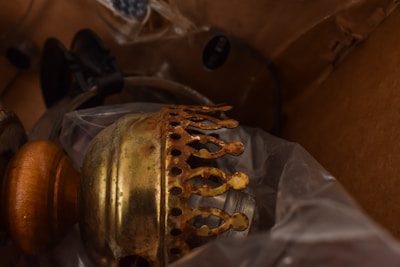 A vintage brass lamp component with a patina finish is surrounded by various other metal and wooden objects. The scene is cluttered, and everything is inside what appears to be a cardboard box. A clear plastic sheet partially covers the items, giving an impression of storage or transportation.