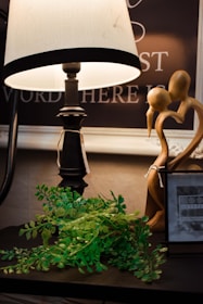 A warmly lit scene featuring a decorative table lamp with a white shade, accompanied by a small green plant with lush leaves. Next to them is a wooden sculpture of two abstract figures intertwined and a photo frame displaying the text 'Sixtrees.' The background includes part of a chalkboard or sign with partial words visible.