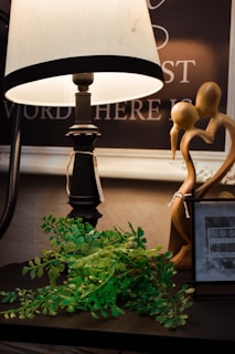 A warmly lit scene featuring a decorative table lamp with a white shade, accompanied by a small green plant with lush leaves. Next to them is a wooden sculpture of two abstract figures intertwined and a photo frame displaying the text 'Sixtrees.' The background includes part of a chalkboard or sign with partial words visible.