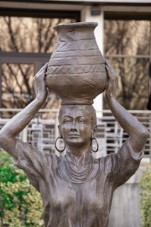 A bronze statue depicts a woman with large hoop earrings and a necklace, balancing a decorated pot on her head. The woman's arms are raised to support the pot, and her expression is serene and composed. The background includes out-of-focus greenery and architectural elements.