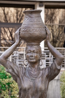 A bronze statue depicts a woman with large hoop earrings and a necklace, balancing a decorated pot on her head. The woman's arms are raised to support the pot, and her expression is serene and composed. The background includes out-of-focus greenery and architectural elements.